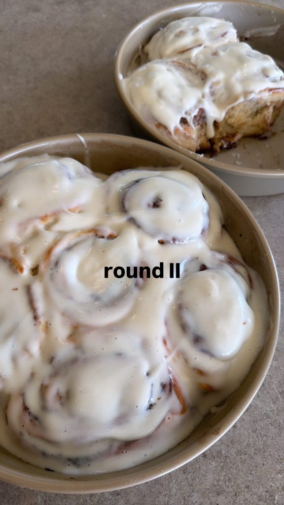 Two baking dishes filled with cinnamon rolls covered in white frosting, with the text "round II" on the foreground dish.