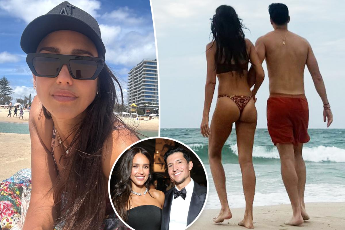 Jessica Alba and Danny Ramirez walk hand-in-hand on a beach in matching red swimwear; Alba wears a cheeky red thong bikini.