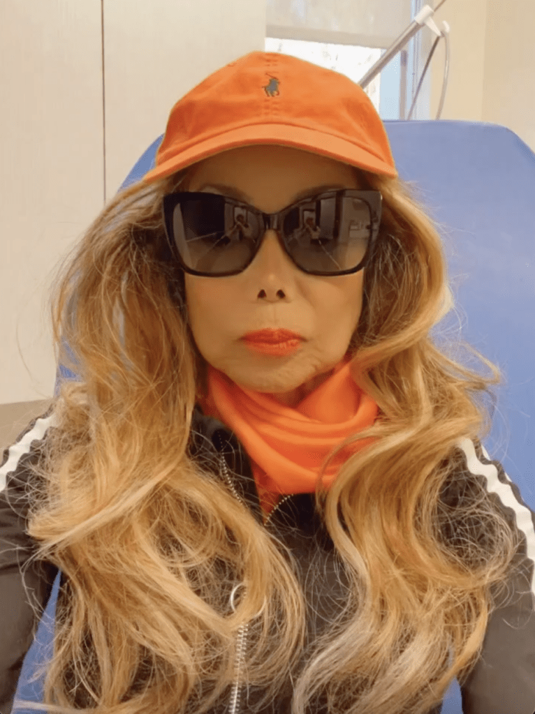 La Toya Jackson wearing an orange hat, black sunglasses, an orange scarf, and a black jacket in what appears to be a doctor's office.