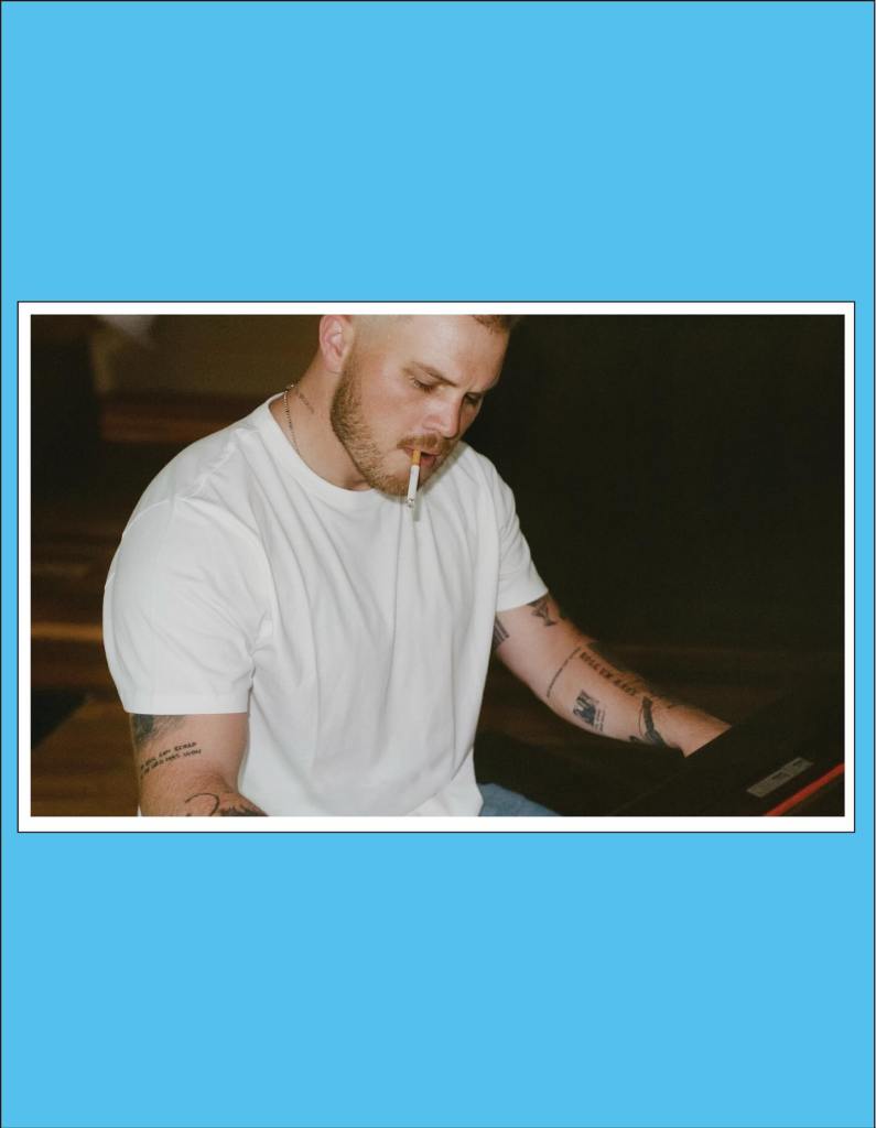 Zach Bryan in a white t-shirt with a cigarette in his mouth, looking down at an object to his right.