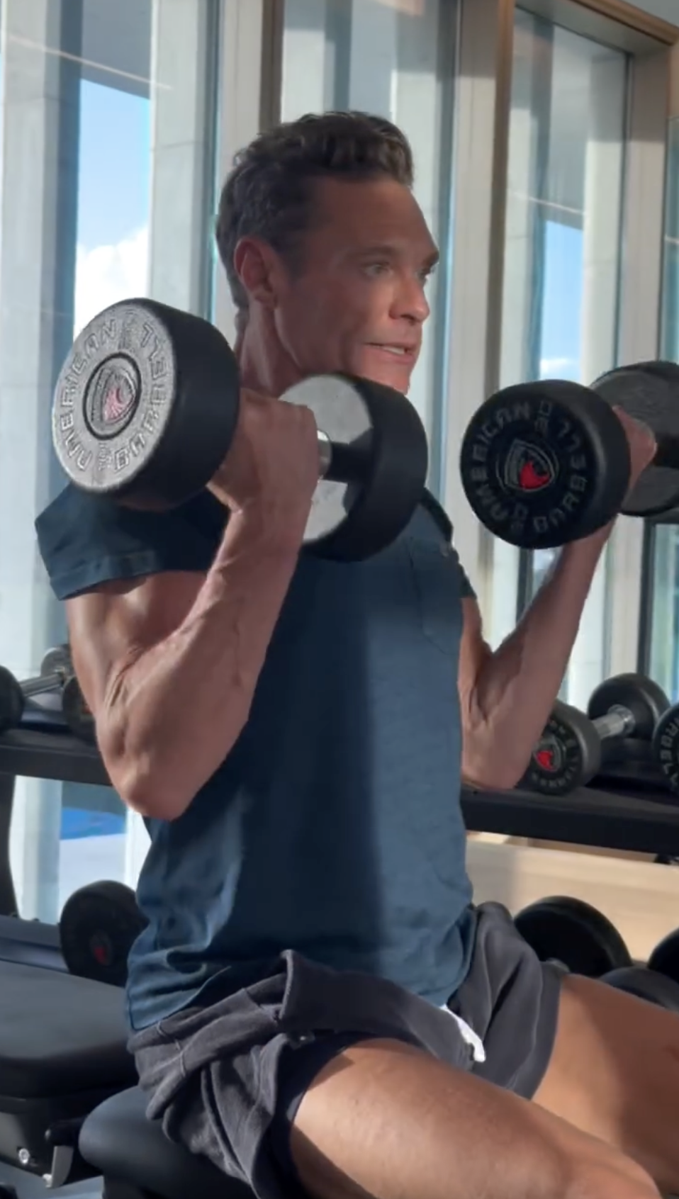 Ryan Seacrest, 50, doing seated bicep curls in a navy T-shirt during a gym workout.