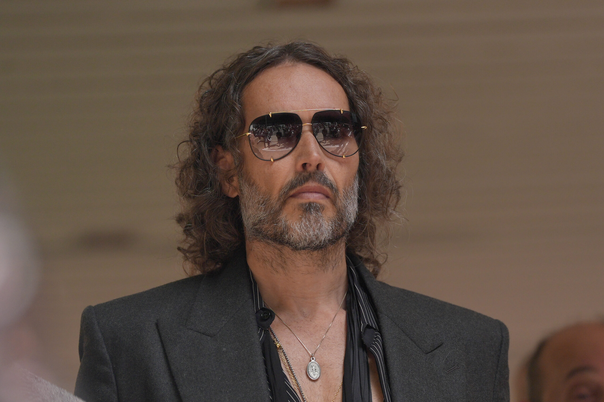 Russell Brand looking forward, wearing sunglasses that reflect a crowd.