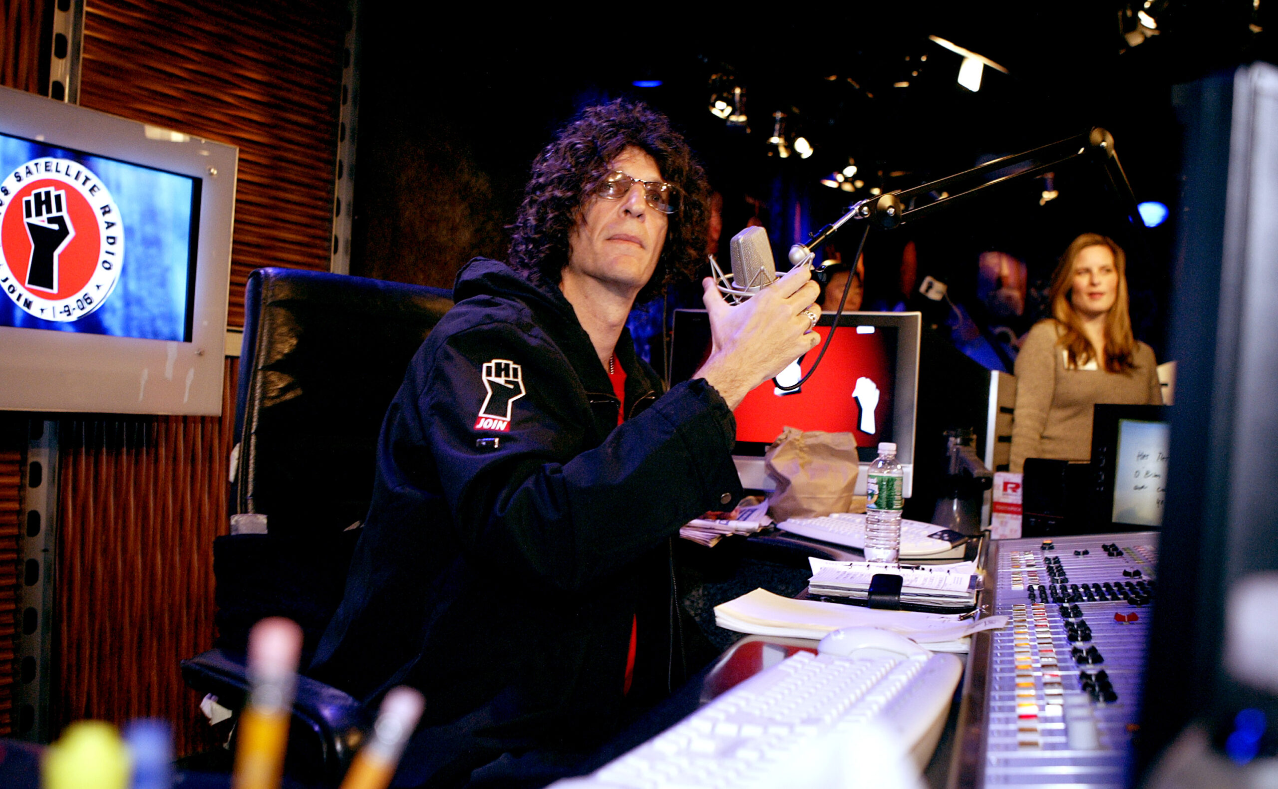 Howard Stern at a Sirius Satellite Radio studio, wearing sunglasses and a black jacket, holding a microphone.