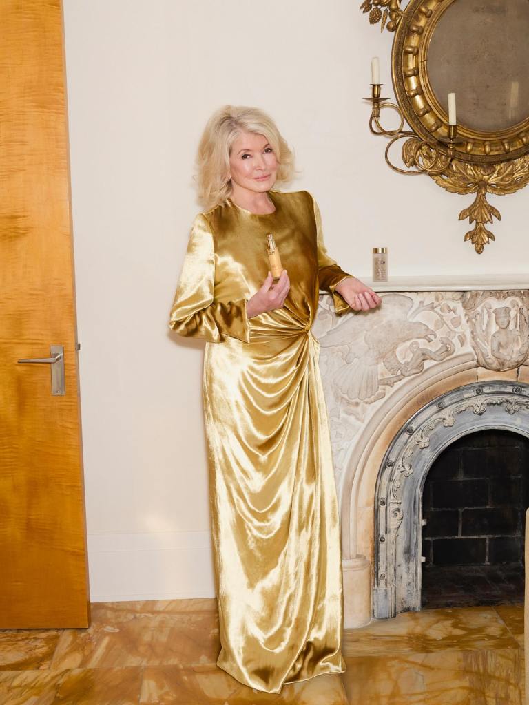 Martha Stewart wearing a gold dress, holding a beauty product, and posing in front of a fireplace.