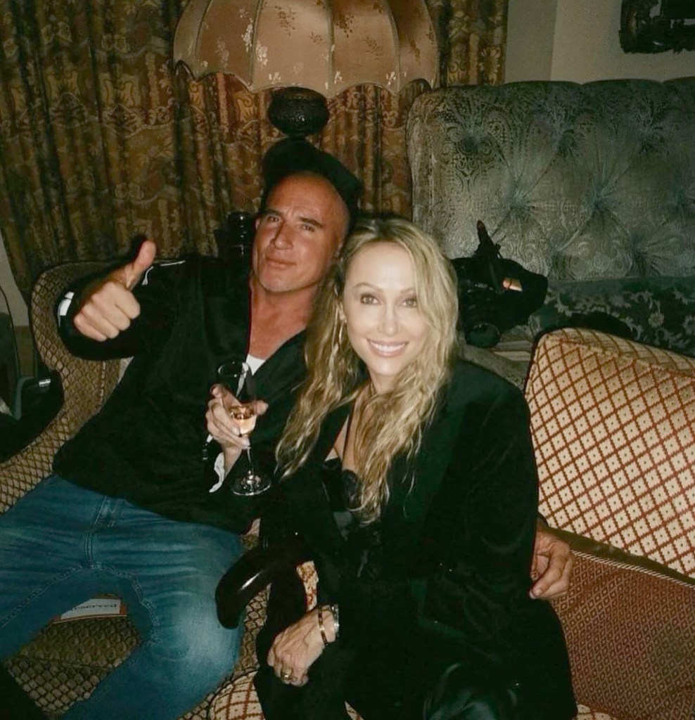 Dominic Purcell and Tish Cyrus sitting on a sofa, with Dominic holding a drink and giving a thumbs up.