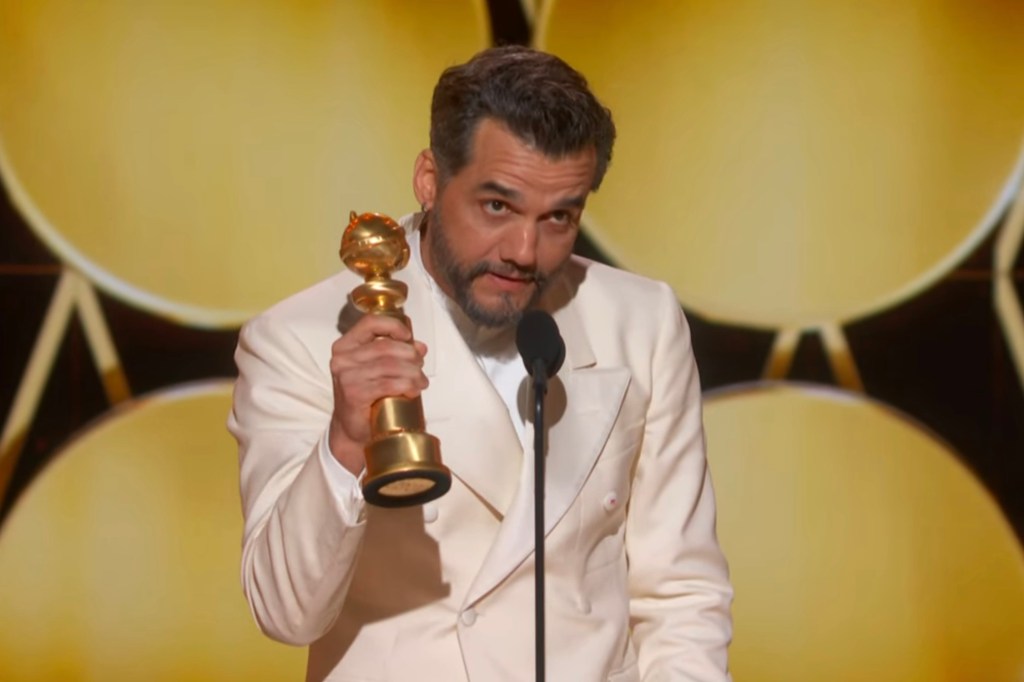 Wagner Moura holds a Golden Globe Award while speaking at a microphone.