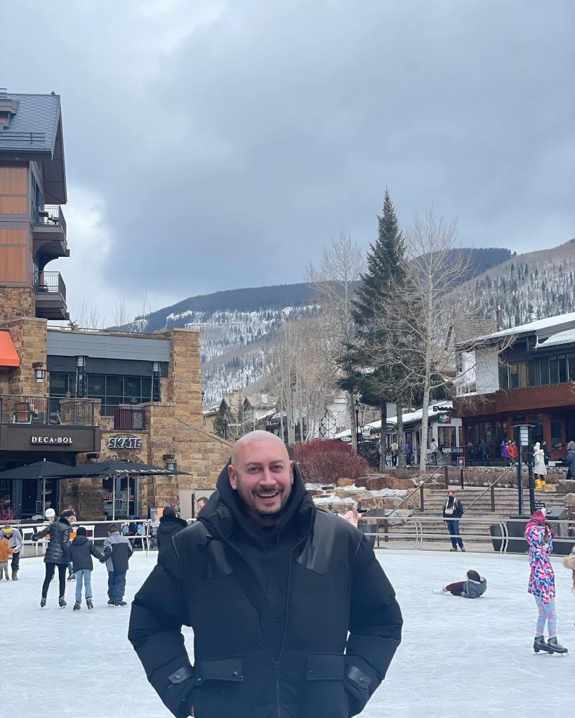 Jasen Kaplan smiling on an outdoor ice rink with snowy mountains in the background.