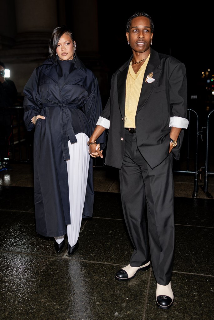 Rihanna wearing a black trench coat over a white dress and A$AP Rocky in a black suit with a yellow knit top, holding hands at the 2025 CFDA Awards.