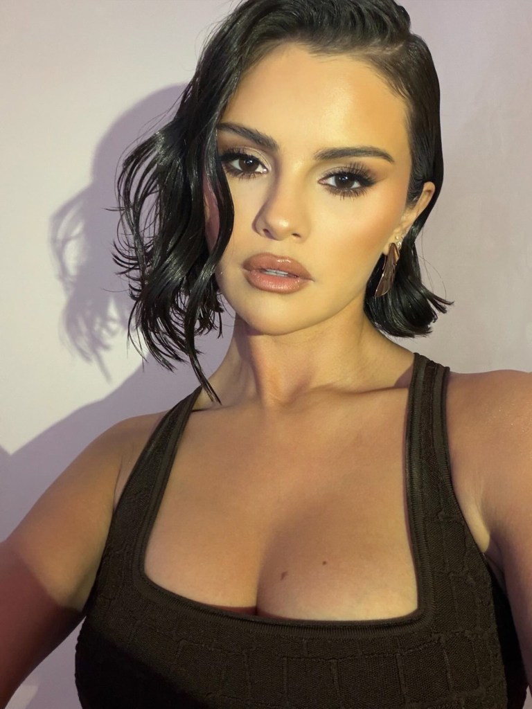 Selena Gomez in full glam with wet-look short curls and feather earrings at a Rare Beauty bronzer event.