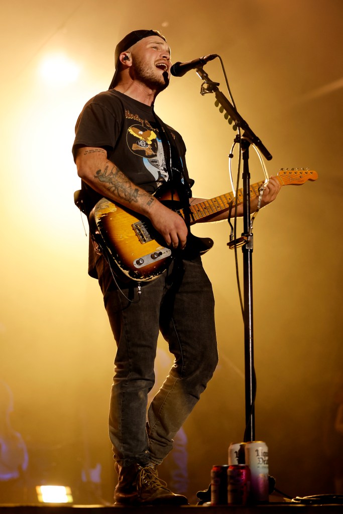 Zach Bryan performing onstage, singing into a microphone and playing a guitar.