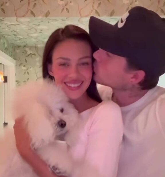 Nicola Peltz holding a white dog while Brooklyn Beckham kisses her cheek.