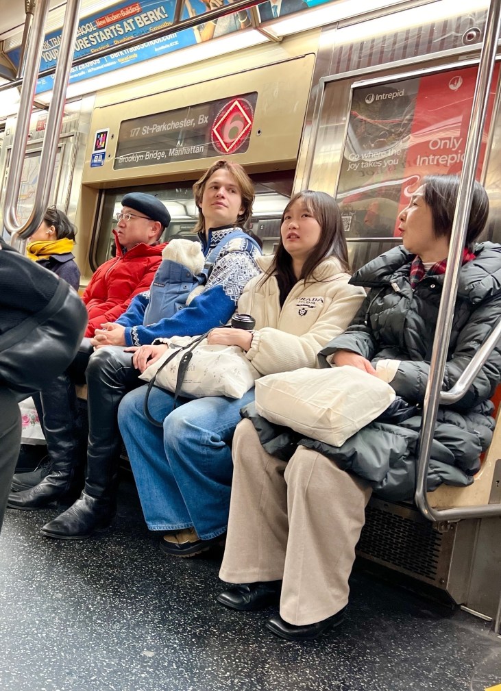 Ansel Elgort rides the New York City subway with a newborn in a carrier; an unidentified woman sits beside him.