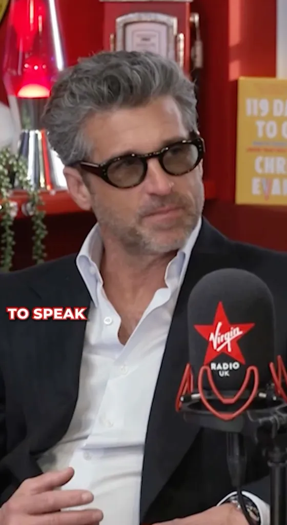 Patrick Dempsey in a dark suit jacket, white shirt, and sunglasses, speaking into a Virgin Radio UK microphone.