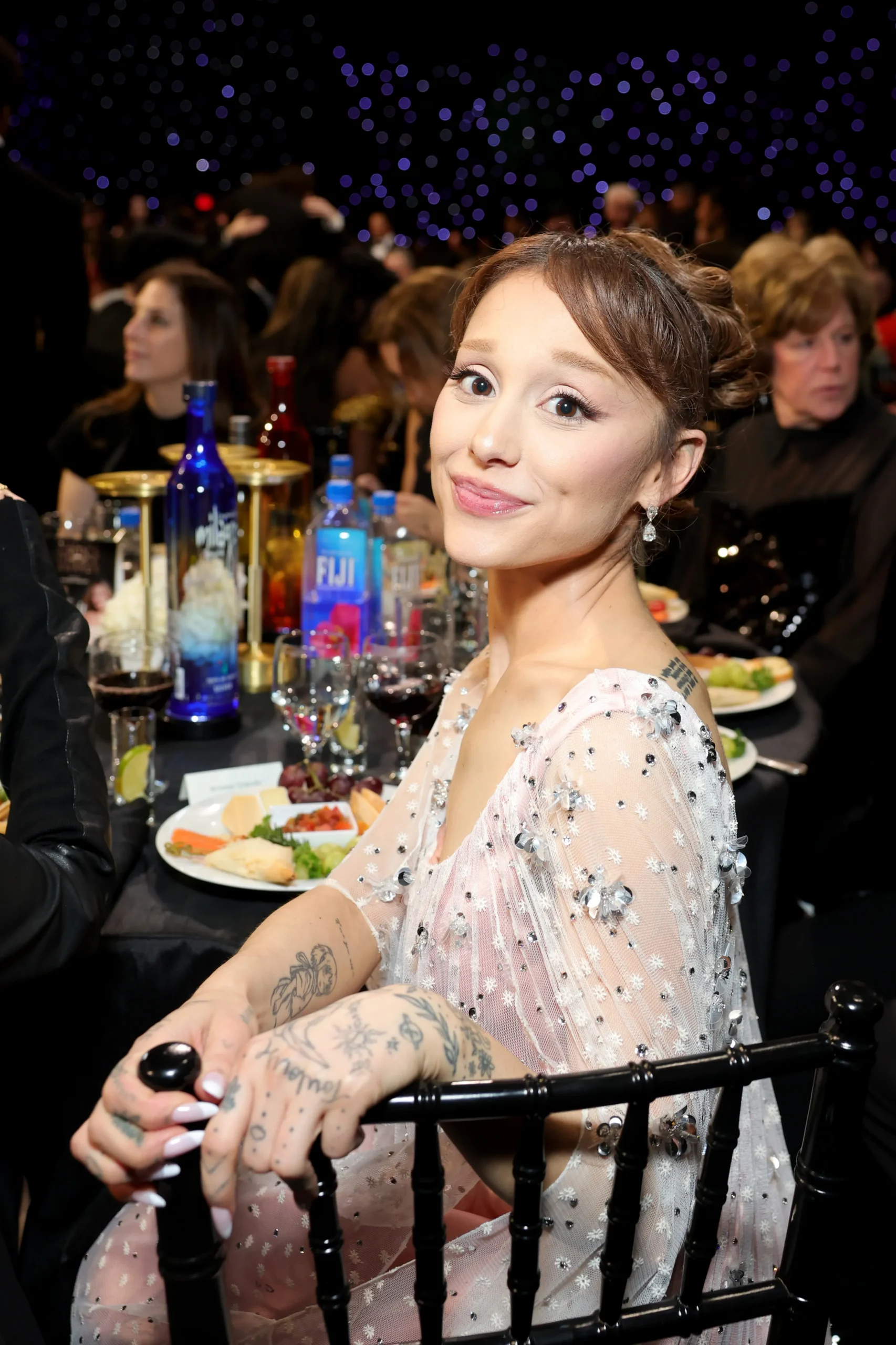 Ariana Grande attends the Critics Choice Awards, wearing a light pink and sheer dress with silver embellishments, smiling at the camera while seated at a table.