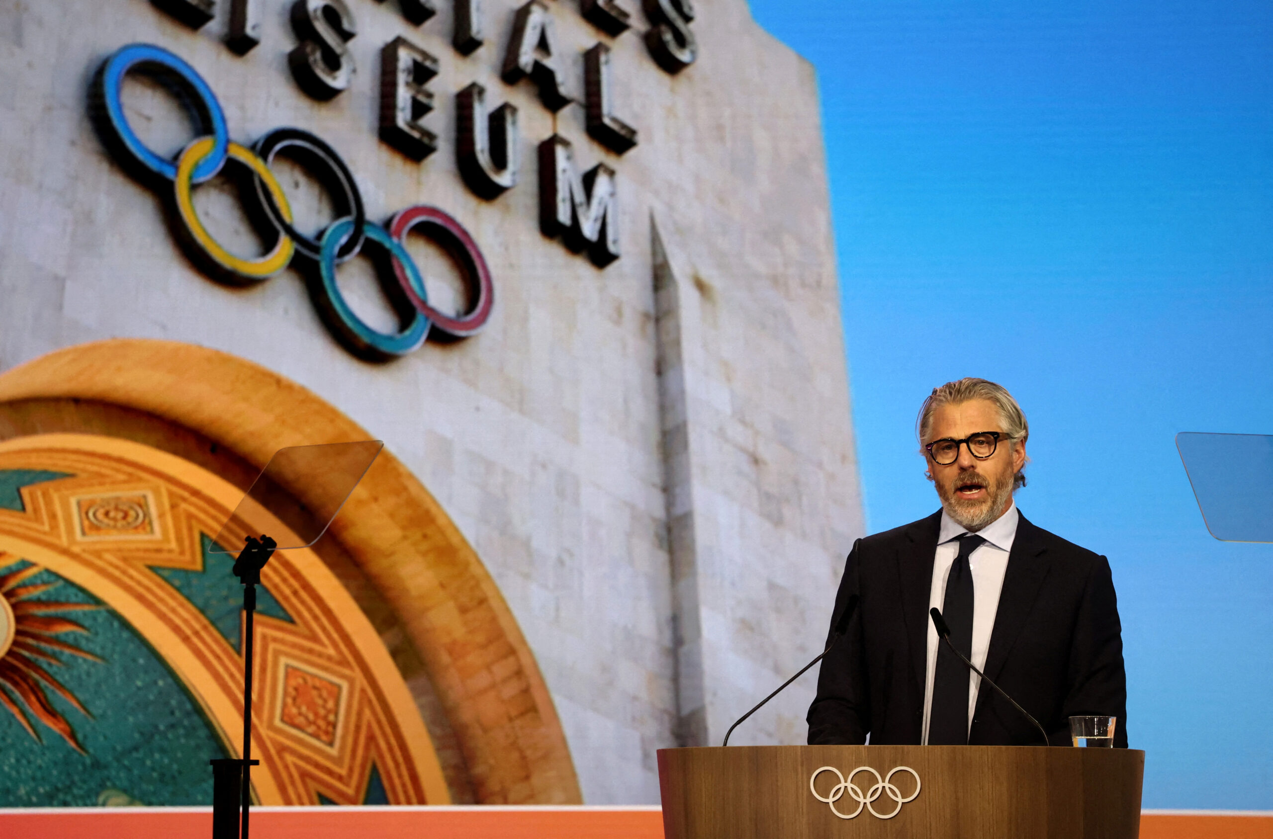 Casey Wasserman addresses the IOC with Olympic rings backdrop in 2025.