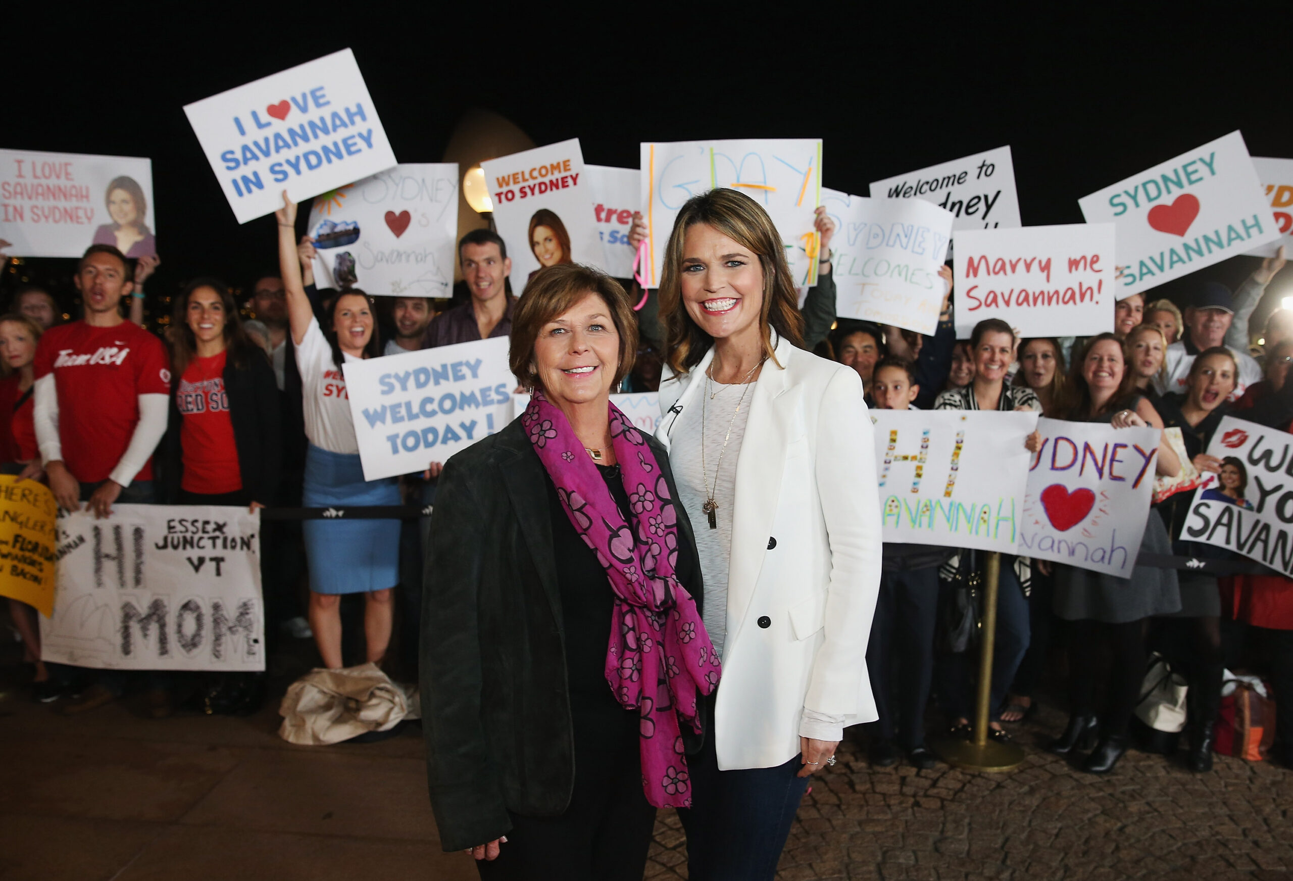 Savannah Guthrie and her mother, Nancy, smiling during a visit to Sydney as a crowd holds welcome signs.