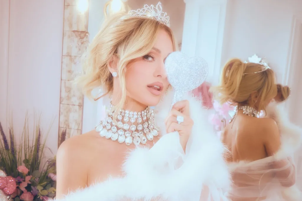 Paris Hilton wearing a tiara and jeweled choker holds a glittery heart-shaped mirror to her eye.