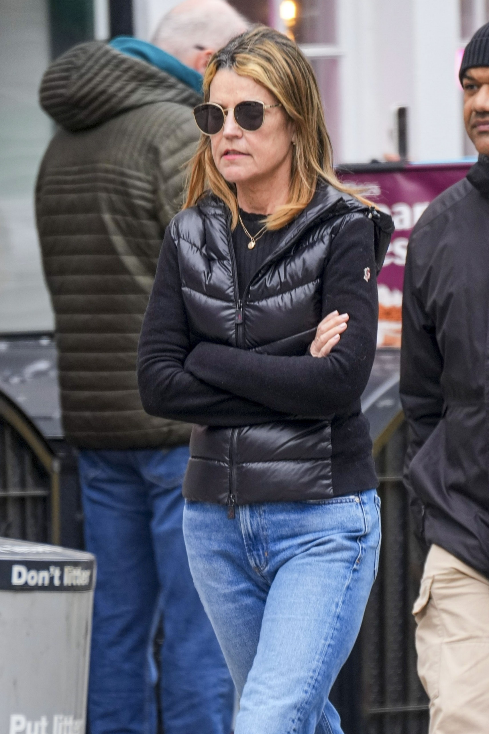 Savannah Guthrie in a black puffer vest, black sweater, blue jeans, and sunglasses, arms crossed, walking in New York.