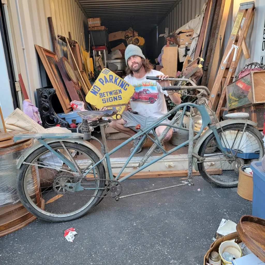 Dusty Riach stands in a storage unit holding a 'NO PARKING BETWEEN SIGNS' sign beside a vintage bicycle.